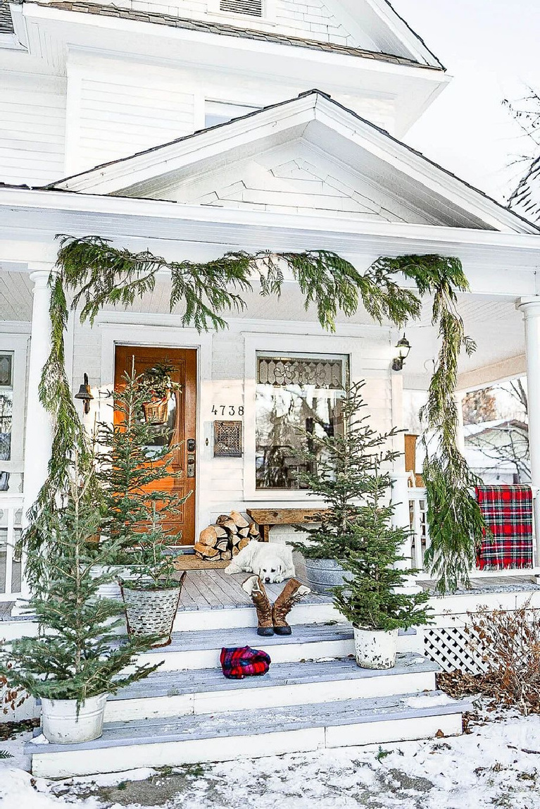 Charming white farmhouse porch decorated simply for Christmas with fresh trees, greenery, and a plaid blanket - Vintage Society Co.