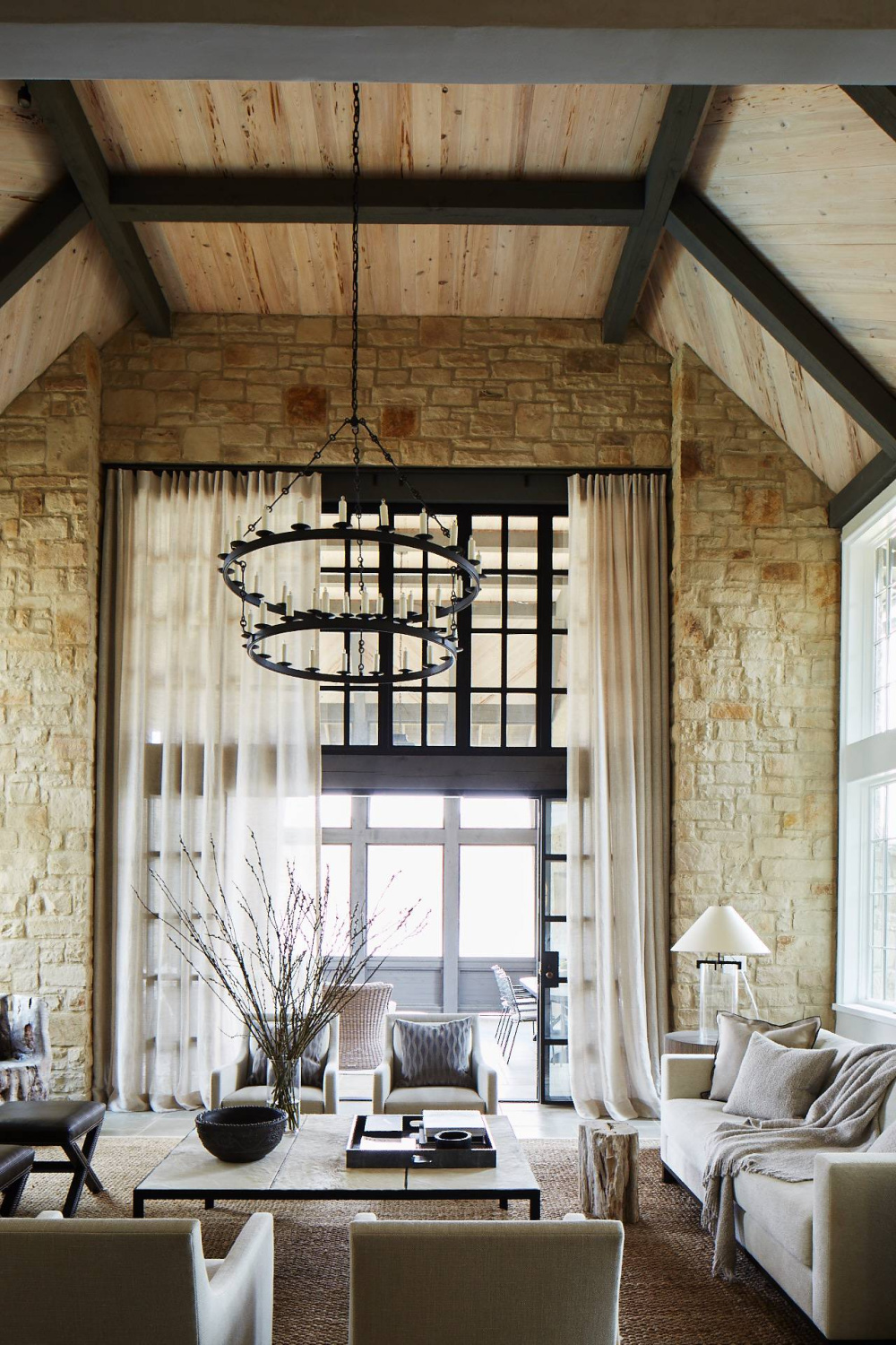 Stunning rustic luxe design in a living room with stone walls, ring style chandeliers, and wood plank ceilings - Jeffrey Dungan.