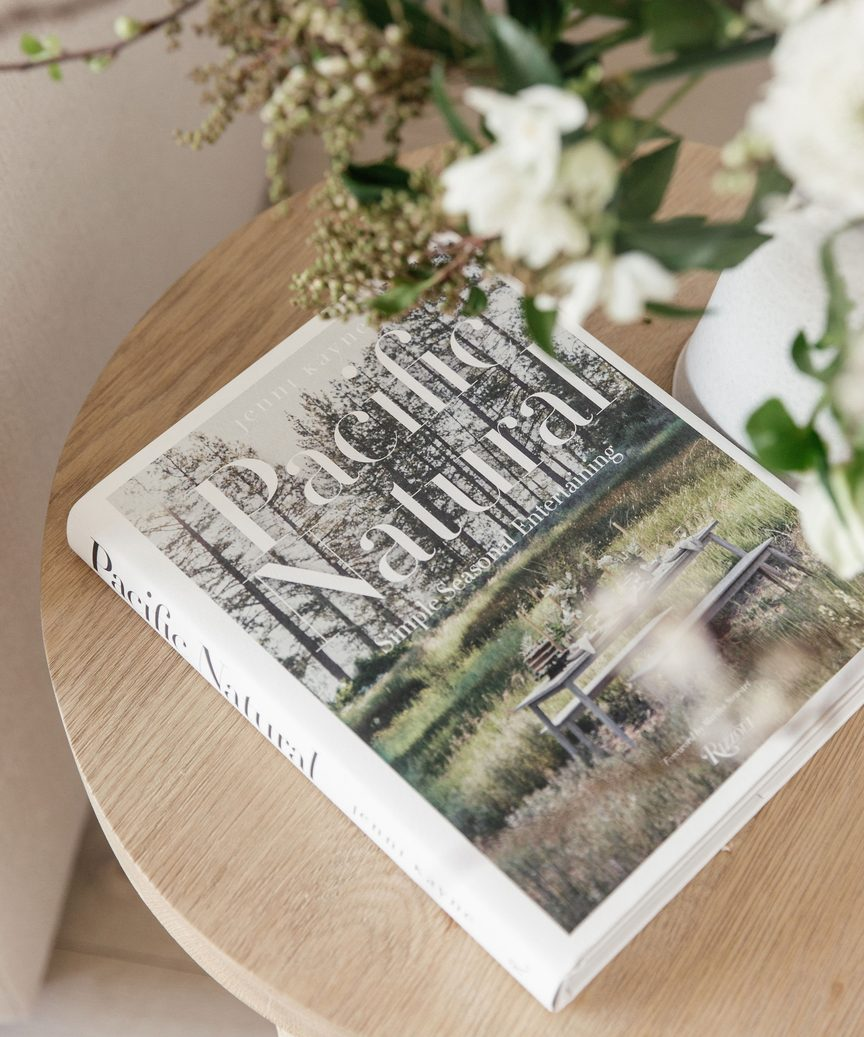 Jenni Kayne's book PACIFIC NATURAL on a light wood round accent table with white floral arrangement.