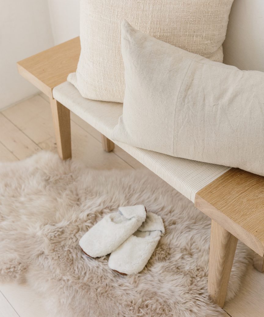 Lovely wood end of bed bench by Jenni Kayne with throw pillows, sheepskin rug, and cozy slippers.