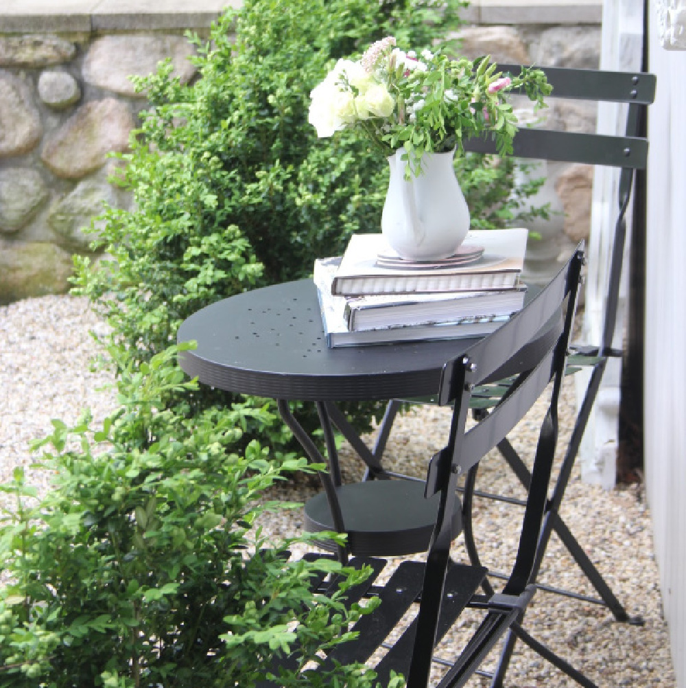 French country style courtyard with pea gravel and stone wall with a black metal modern French bistro dining and boxwood. #hellolovelystudio #frenchcountry #modernfrench #cafechairs #outdoordining #outdoorfurniture #patiofurniture