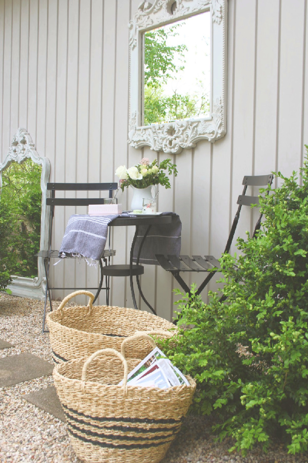 French country courtyard with boxwood, gravel, black bistro set, baskets, and a French mirror. #hellolovelystudio #cafechairs #frenchcountry #bistrochairs #outdoordining #frenchbaskets #patiofurniture #outdooroasis