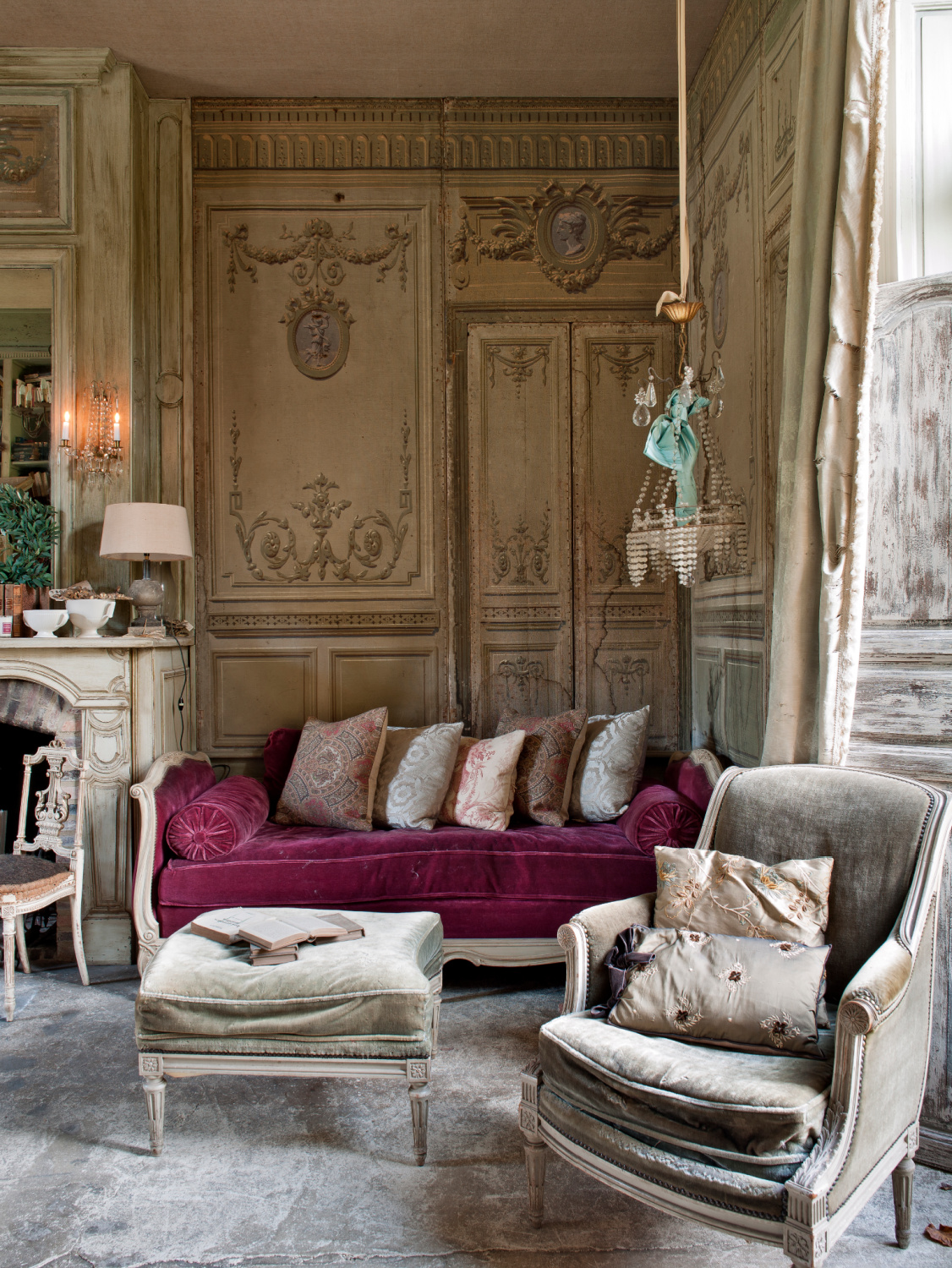 Traditional French provincial sitting room with painted walls and, unusually, a concrete floor. A sofa with a maroon red cushion stands in one corner. Featured in Shauna Varvel's PROVENCE STYLE. #interiordesign #frenchcountry #oldworldstyle #velvet