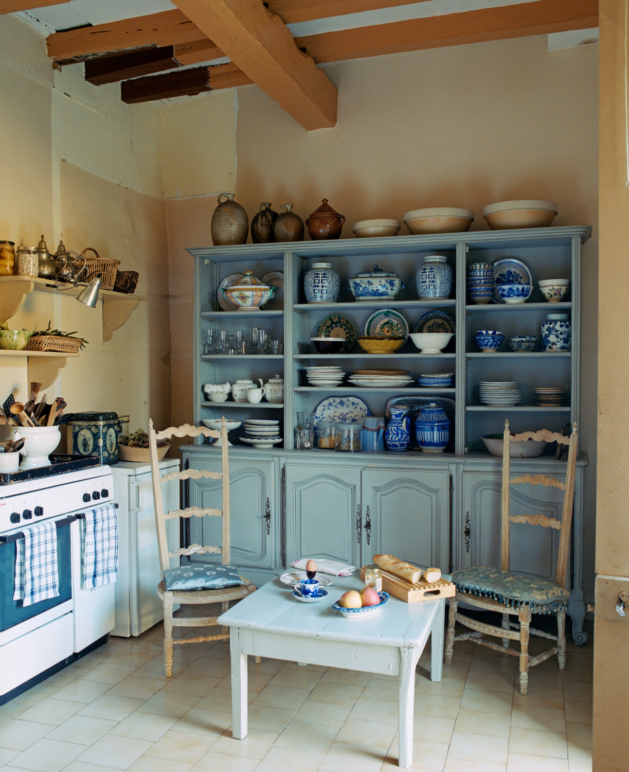 A pair of high-backed kitchen chairs in front of large blue dresser in French provincial kitchen displaying a collection of ceramics from North Africa, Andalucia, China and Holland. Featured in Shauna Varvel's PROVENCE STYLE. #interiordesign #frenchcountry #oldworldstyle