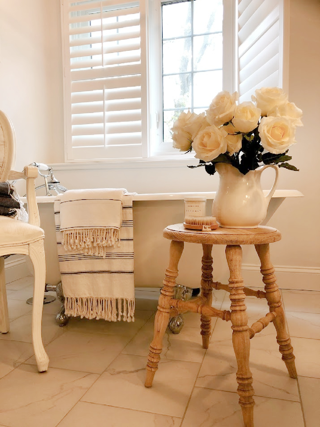 French country white bathroom with maids tub, accent stool, and white roses - Hello Lovely.