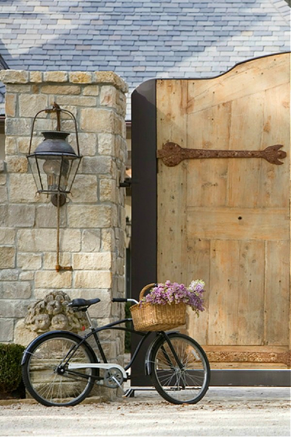 Charming French country rustic entry with wood gate, black bicycle with basket of blooms, and rugged stone and lantern - Chateau Domingue.