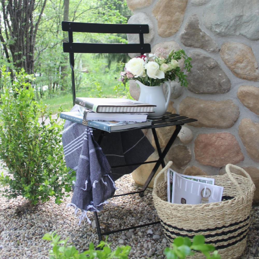 French bistro chair and stripe Turkish throw tucked in a corner of my garden to invite relaxation and reading (cookbooks are my idea of relaxation!). #hellolovelystudio #frenchcountry #bistrochair #turkishthrow #seagrassbasket #turkishtowel #outdooroasis #romanticgarden #outdoordecor #bistrochairs