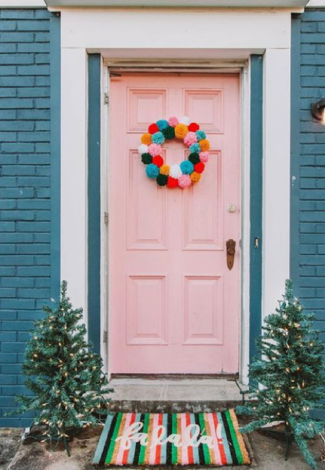 Colorful boho pompom wreath on pink door of blue brick house with striped falala doormat. #christmaswreath #pompomwreath #pinkchristmas