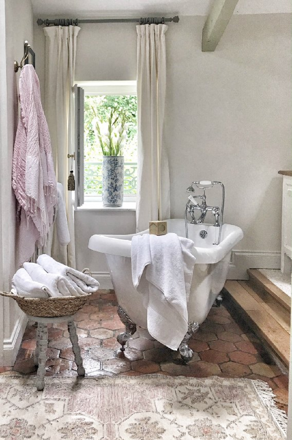 Quiet and timeless hues in a French farmhouse bathroom by Vivi et Margot features reclaimed terracotta hex tiles, freestanding tub, romantic chandelier, and Farrow and Ball's Strong White on the walls.