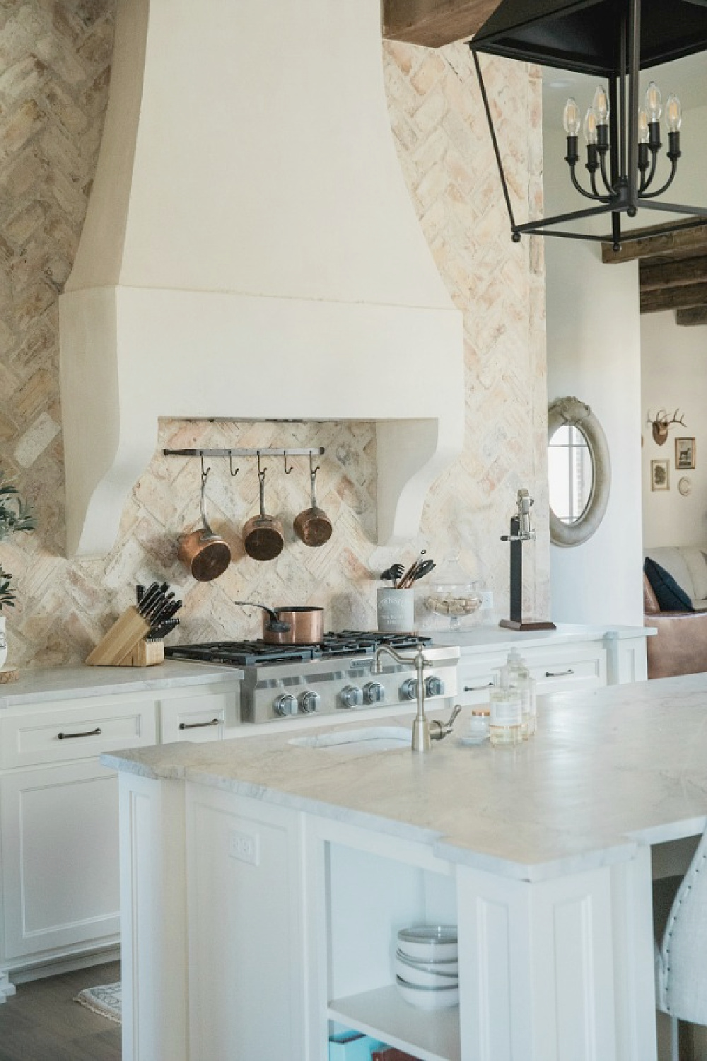 Chicago brick backsplash and custom French country range hood with copper pots on rack beneath in a country French kitchen by Brit Jones Design.