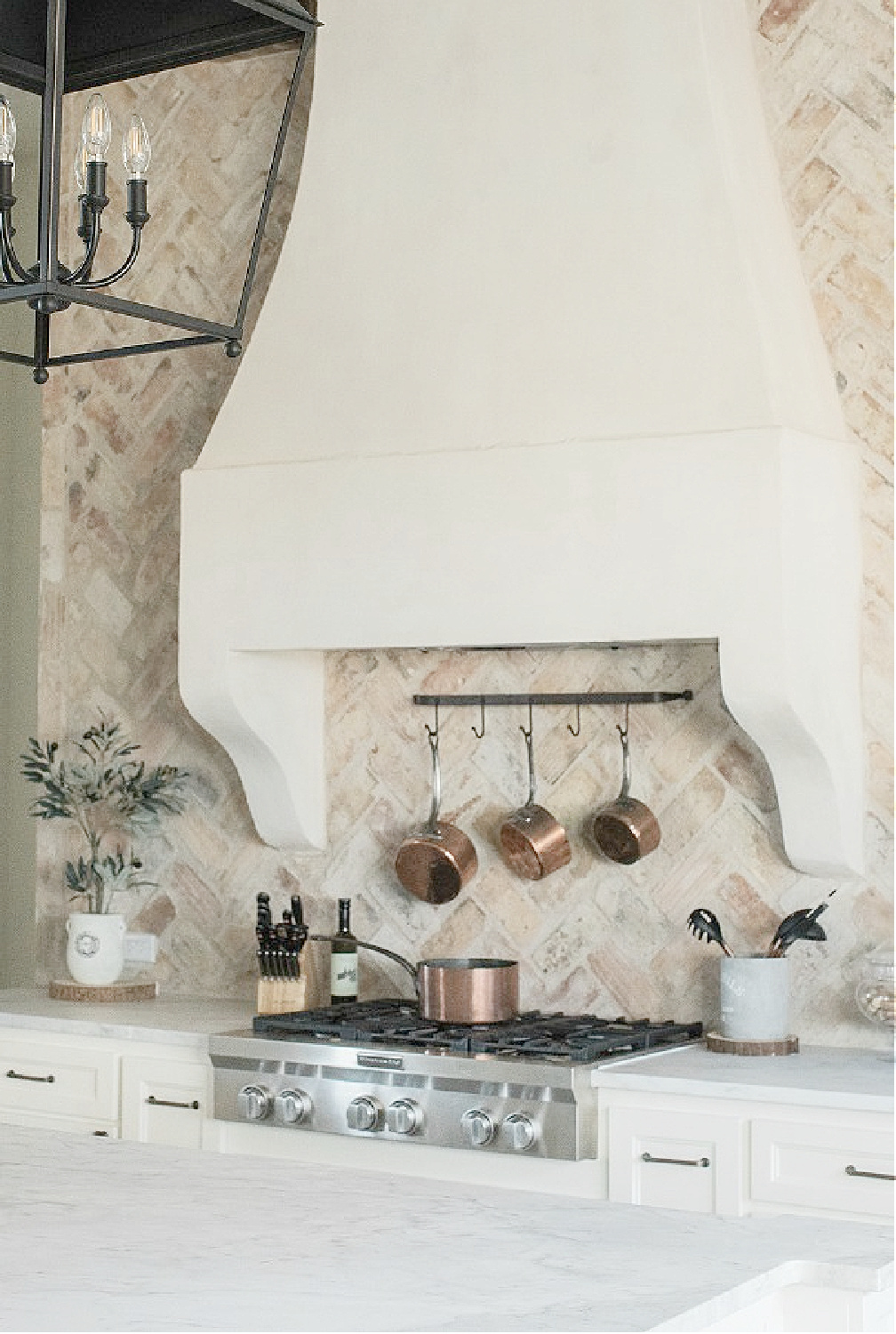 Chicago brick backsplash and custom French country range hood with copper pots on rack beneath in a country French kitchen by Brit Jones Design.