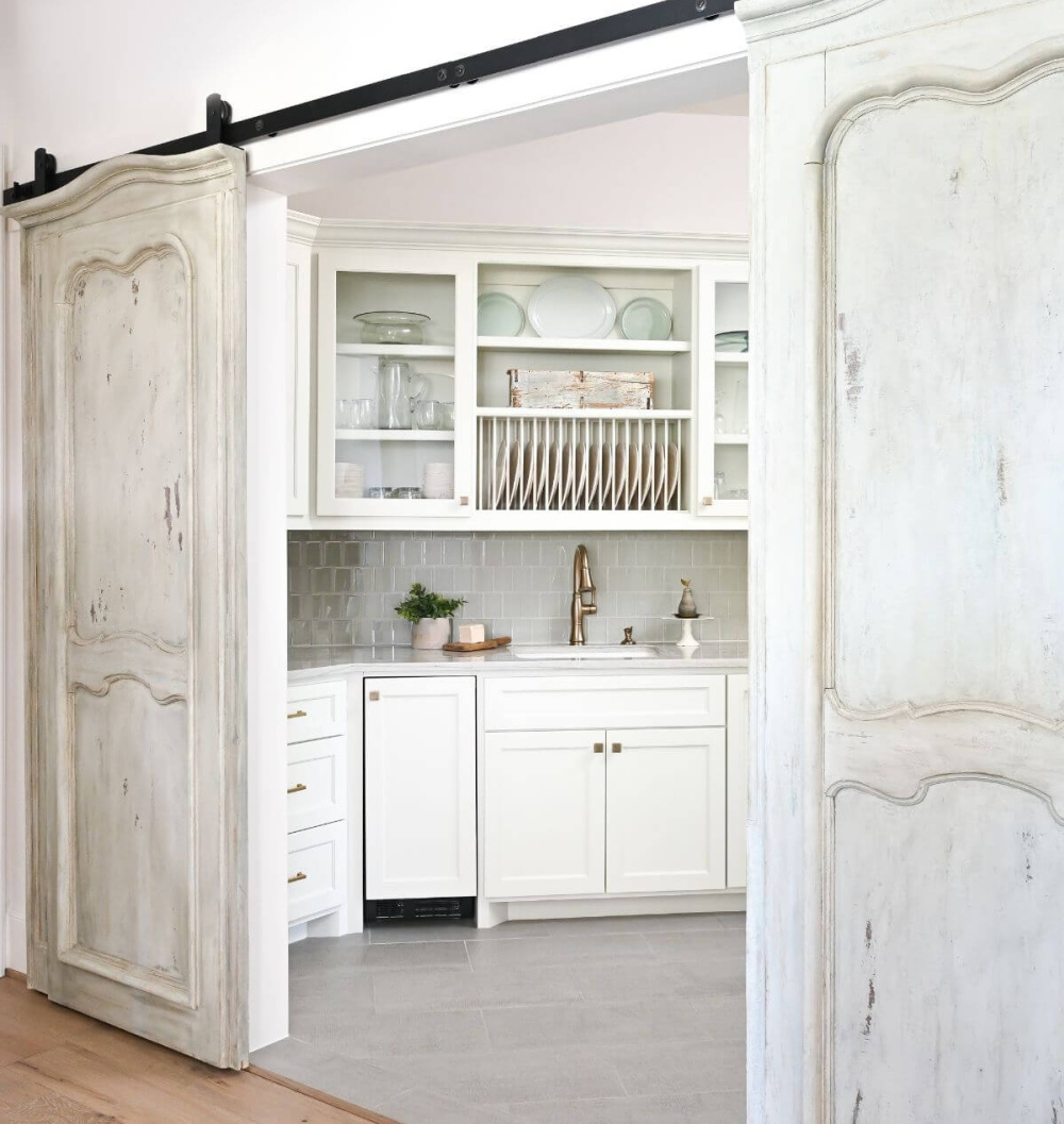 Beautiful white pantry with antiqued barn doors in a French country kitchen - Morningstar Builders. #frenchcountrypantry #frenchkitchen #pantrydesign