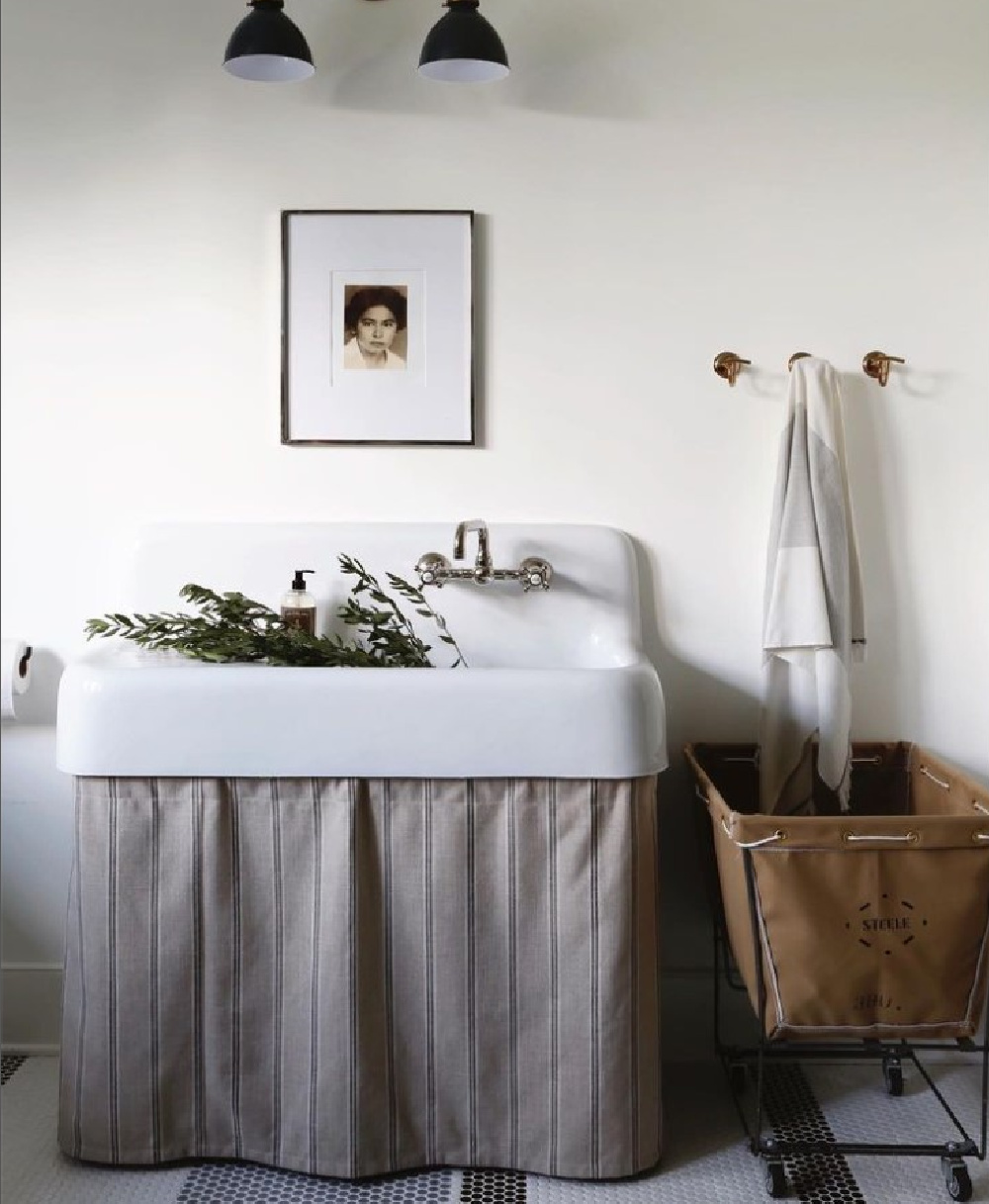 Minimal, serene laundry room with vintage farm sink with skirt, laundry cart, and black and white tiled classic flooring - design by April Tomlin Interiors. #laundryroomgoals #traditionalstyle