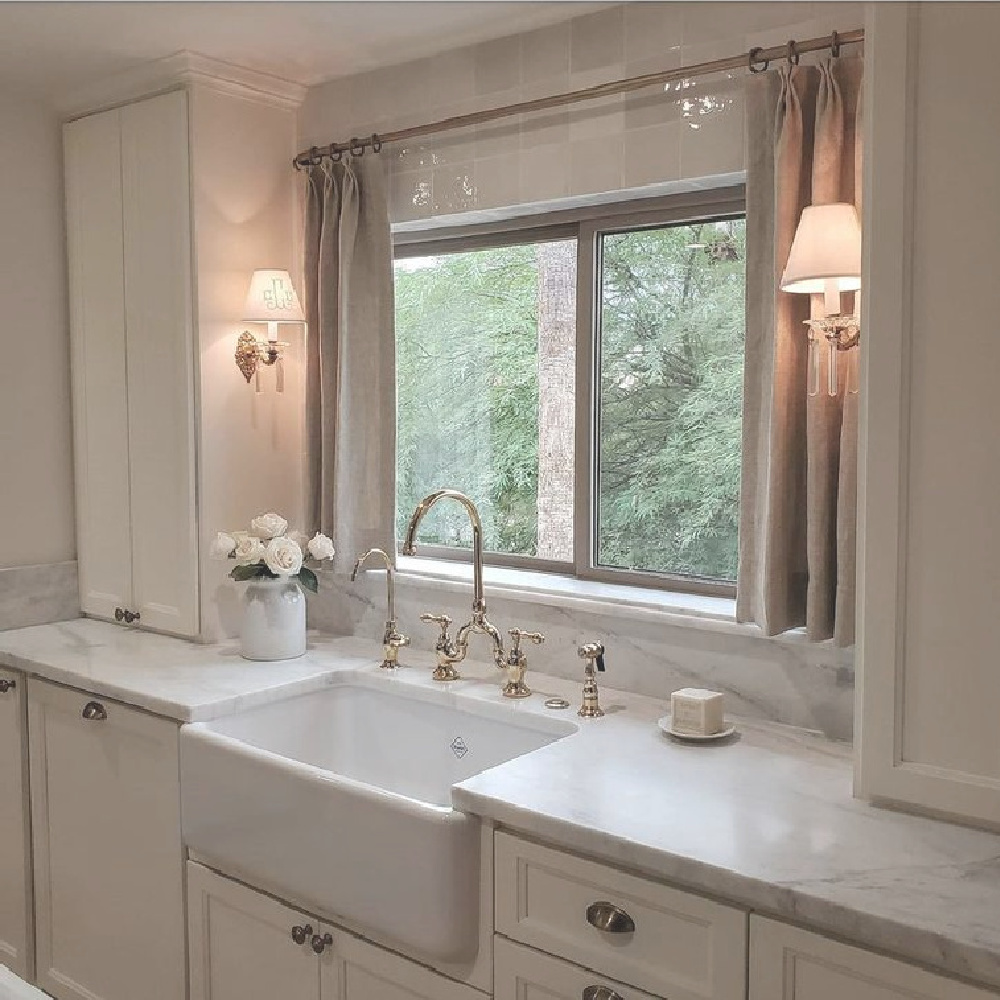 Romantic all white French country kitchen by French Nest Interior Design Co with farm sink flanked by sconces, linen curtain panels, and simple classic cabinetry. #frenchcountry #kitchens #frenchkitchens #kitchendesign #farmsink #romanticdecor