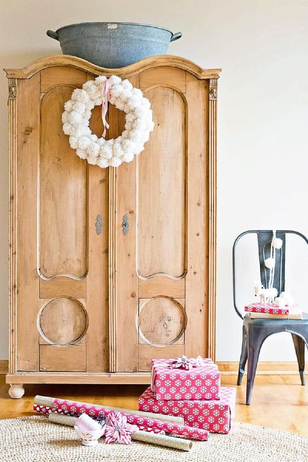 Farmhouse Christmas decor with a scrubbed wood armoire with arched doors, fluffy pom pom wreath, and gifts wrapped in pink - A Rosy Note.