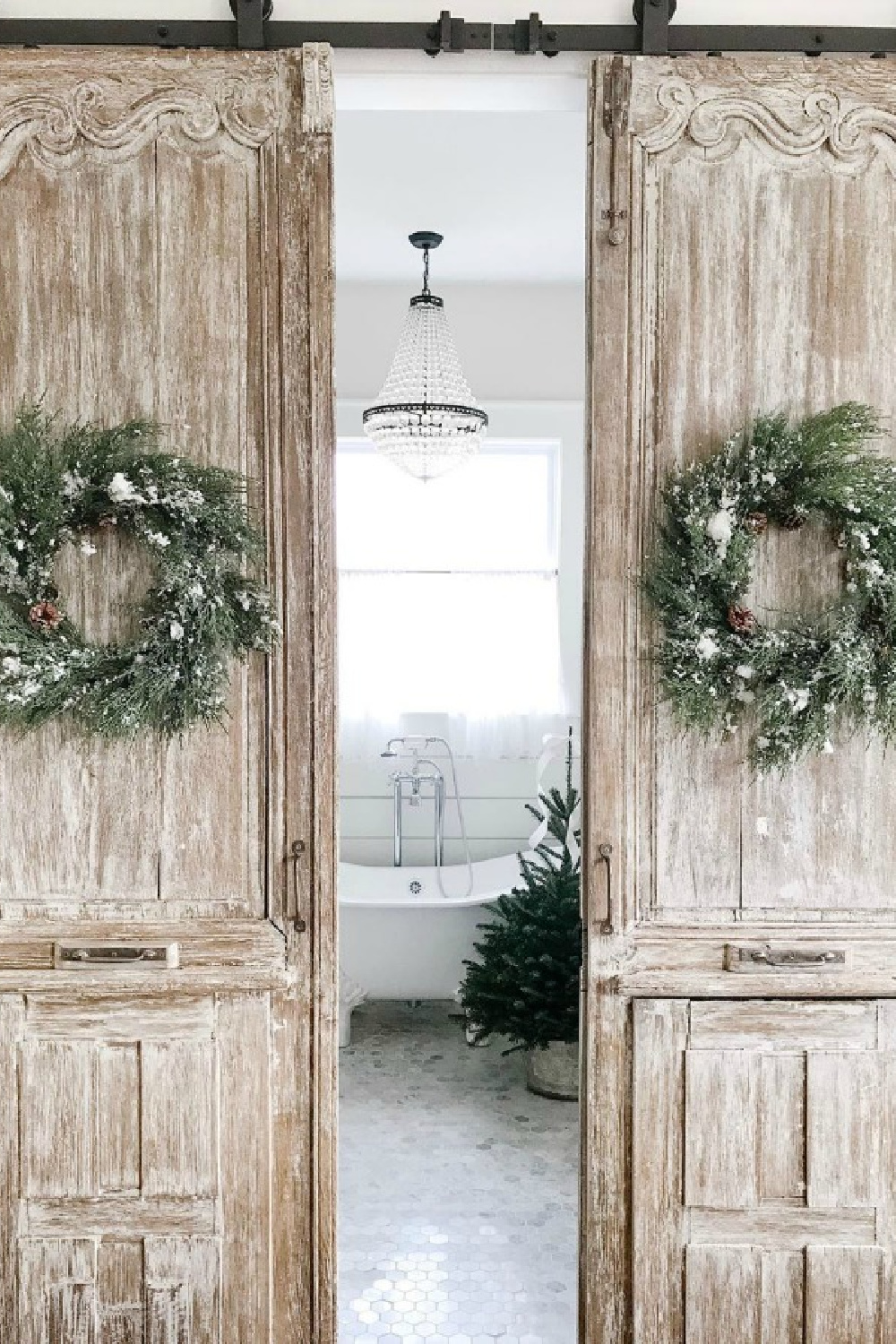 Sliding barn doors decorated with Christmas wreaths and open to a lovely bath with empire crystal chandelier over clawfoot tub - Beside the Mulberry Tree. #farmhousechristmas #christmasdecor #rusticchristmas