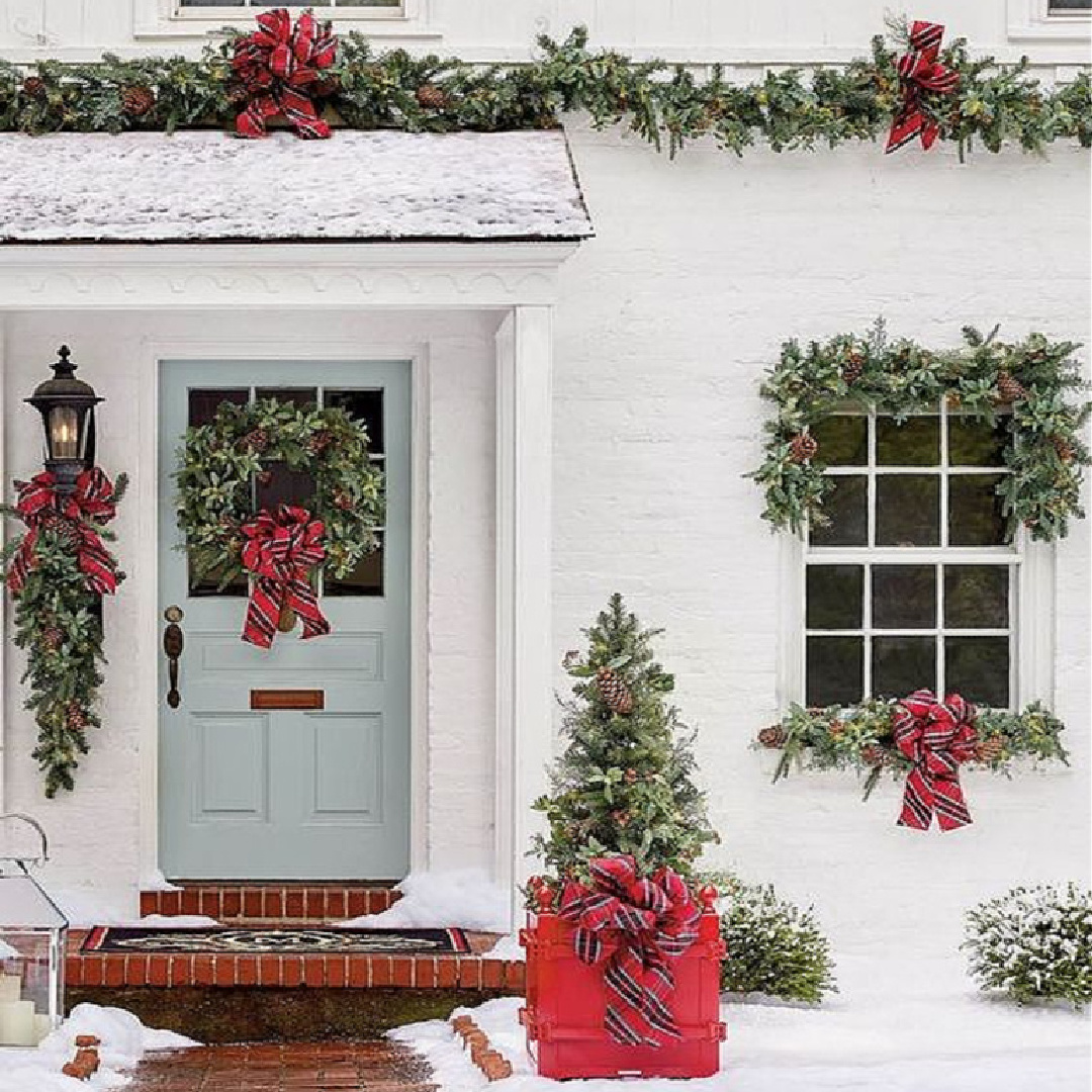 Charming white brick exterior of a country home decorated for Christmas with greenery and plaid ribbon - Frontgate. And that light blue door!