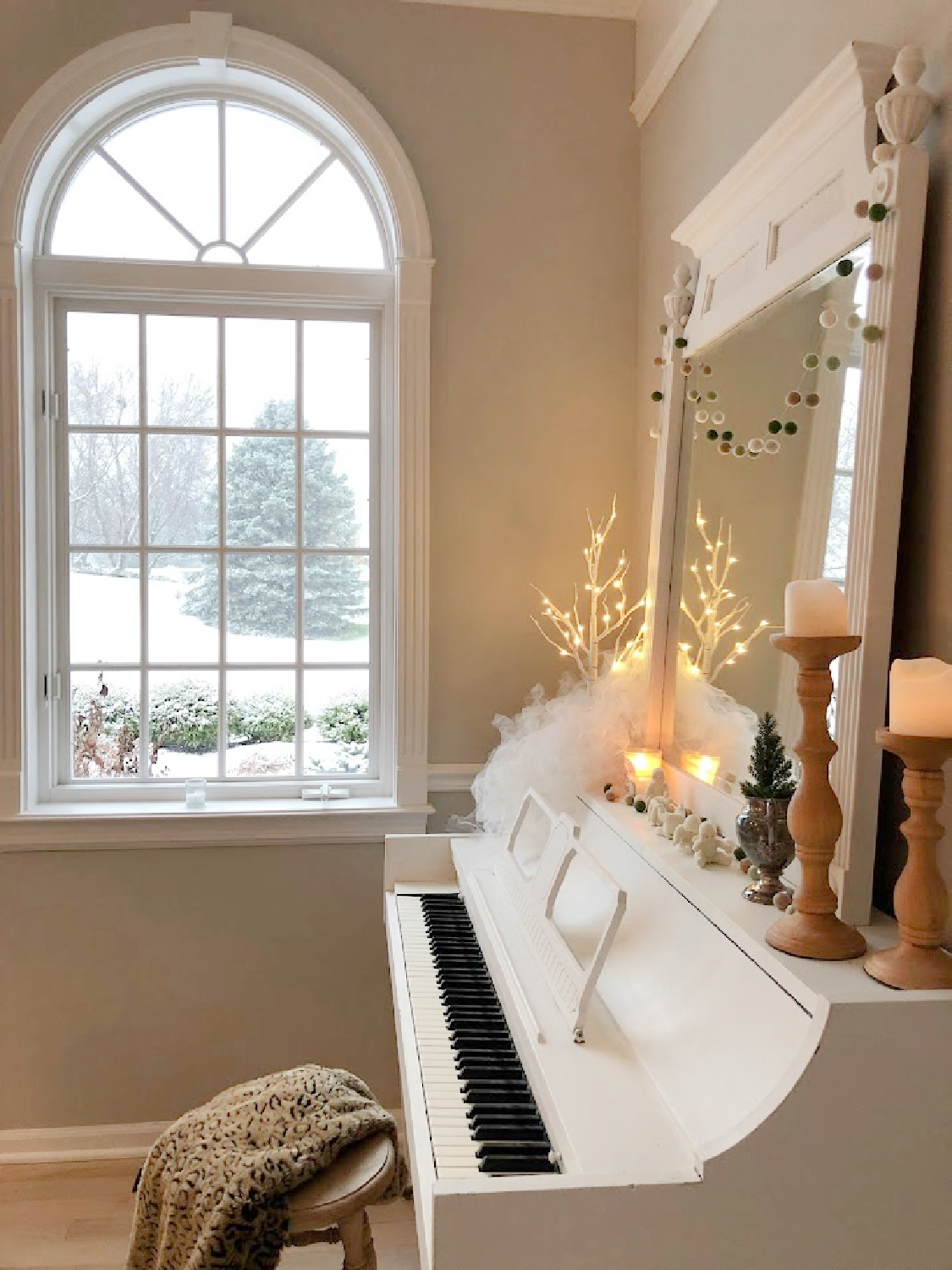 My vintage white piano decorated for Christmas and snow outside the arched window in the music room - Hello Lovely Studio.