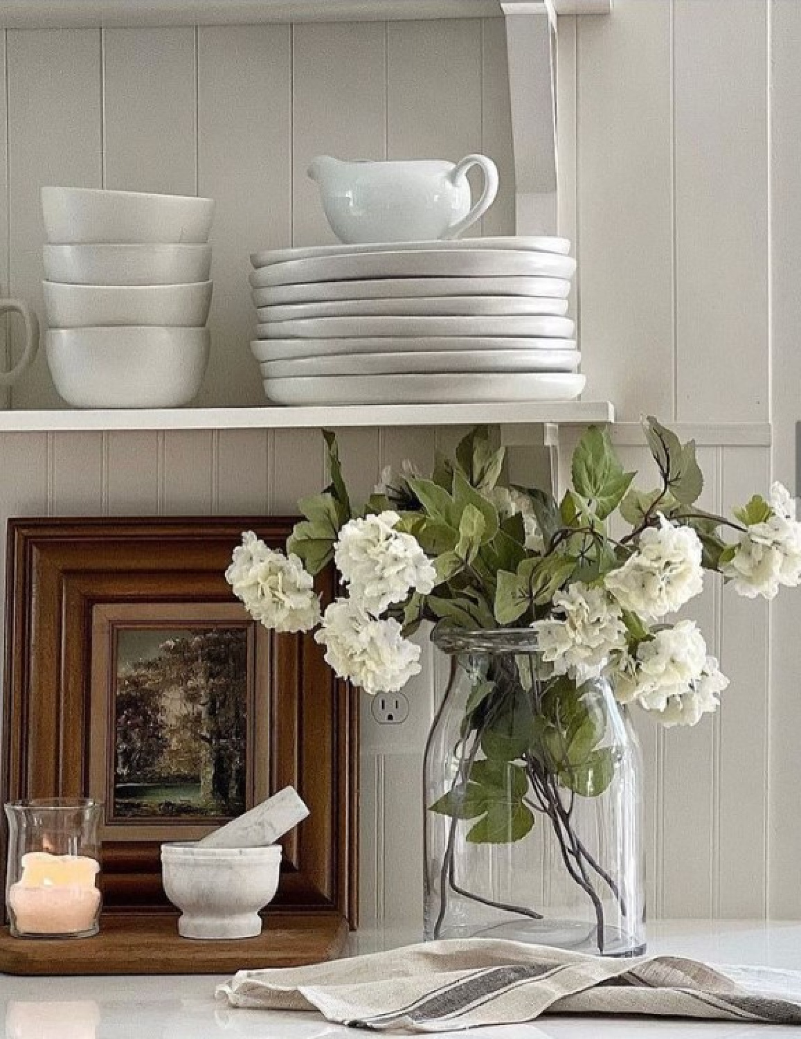 Beautiful American country farmhouse vignette in a white kitchen with open shelving stacked with plates, white flowers in simple jar, mortar and pestle and cande - @handmade_farmhouse. #americancountry #whitecountrykitchen