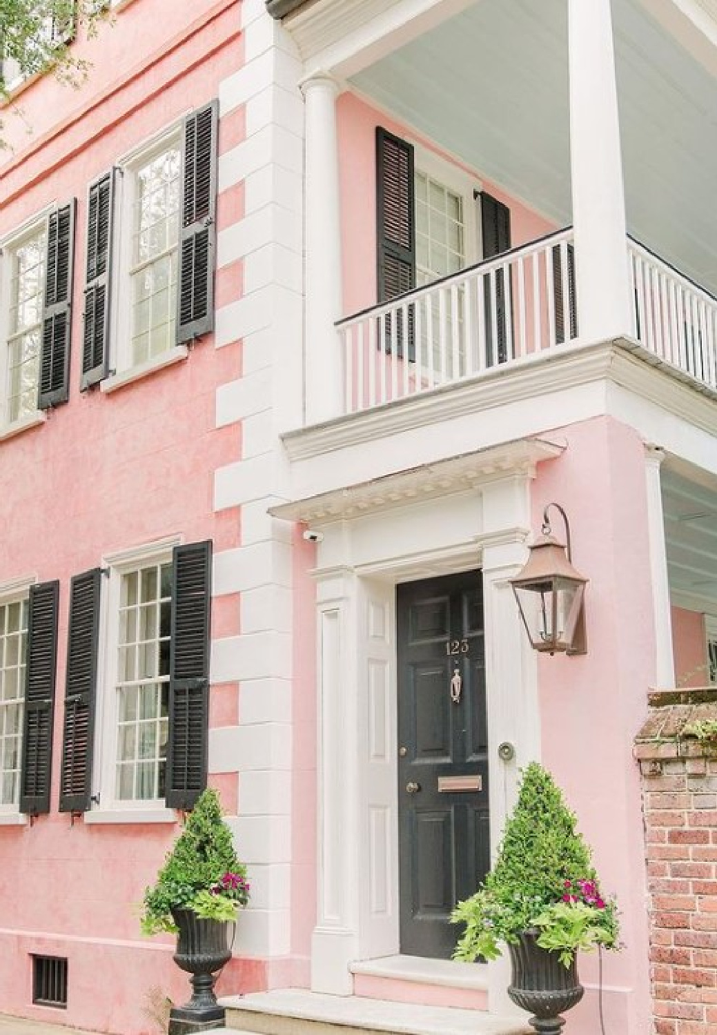 Pink Charleston house exterior with black shutters and front door, topiaries, and gas lanterns - @palmbeachlately (photo by SarahLainePhoto). #pinkhouses #charlestonhouses #pinkpaintcolors