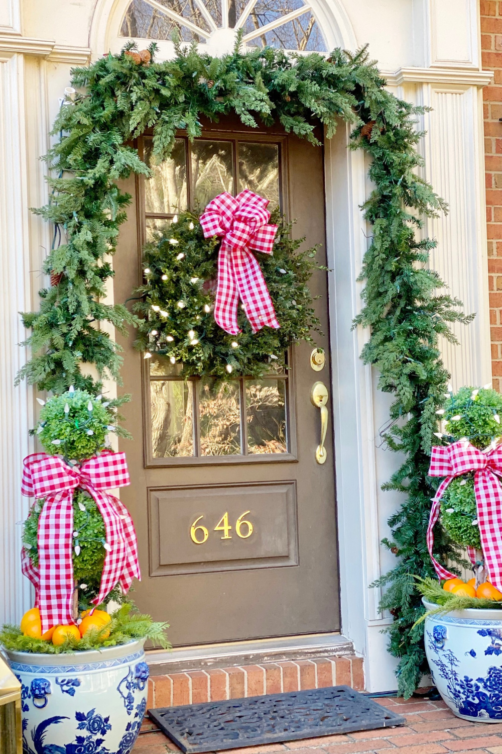 Preppy pink check ribbon on a Christmas porch with garland, blue porcelain topiaries, and a sweet wreath on the door - The Pink Clutch Blog. #pinkchristmas #preppychristmas #christmasporch