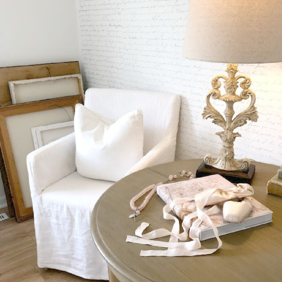 Pink pointe shoes on a round table in a bedroom with white linen slipcovered chair, script stenciled wall, and canvases on floor - Hello Lovely Studio.