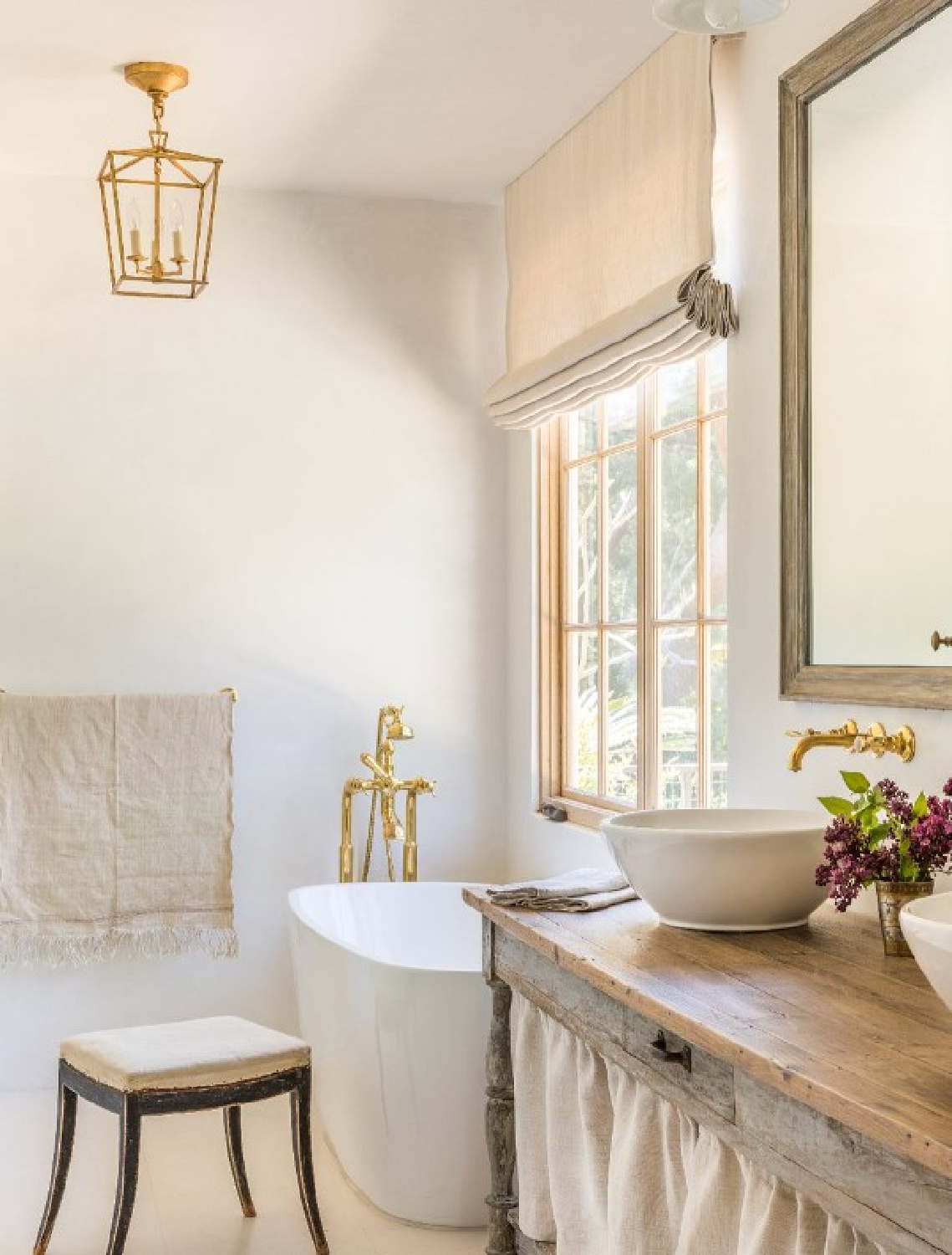 Giannetti Home designed timeless European country inspired bathroom with antique vanity, wall mount unlacquered brass faucet, Darlana mini gilded lantern, and soaking tub.