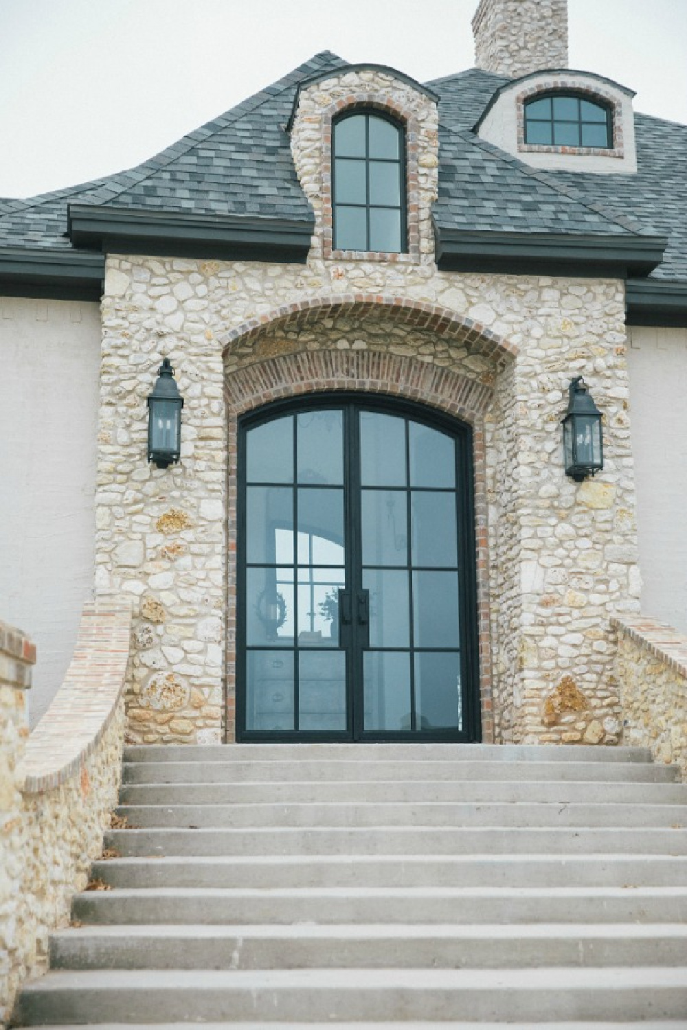 Elegant French country home esterior with steel double arch front doors, stone, brick, stucco, and rustic lanterns. Design by Brit Jones.