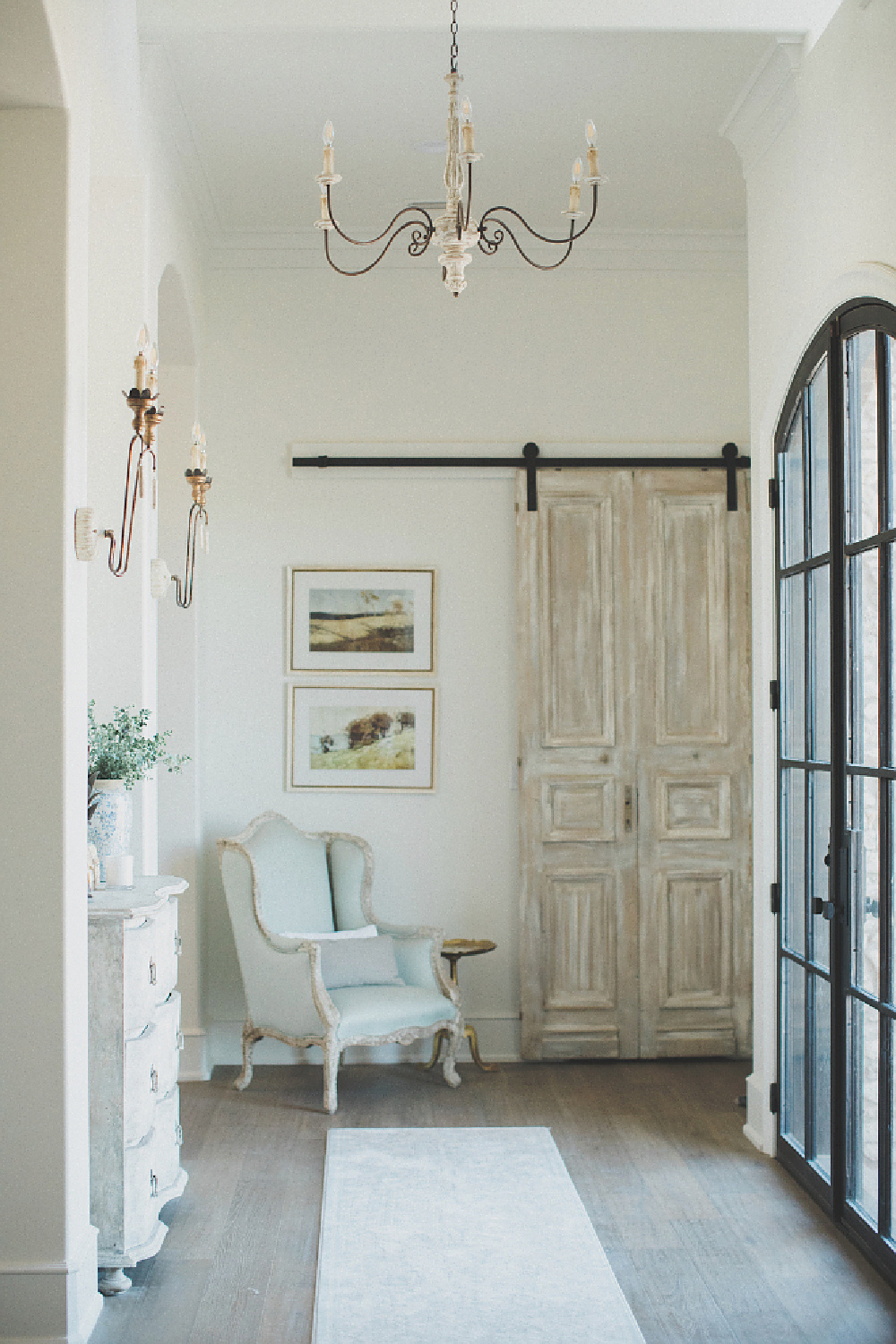 French country farmhouse entry with rustic barn door, wingback chair, steel windows, chandelier and wall sconces. Brit Jones Design.