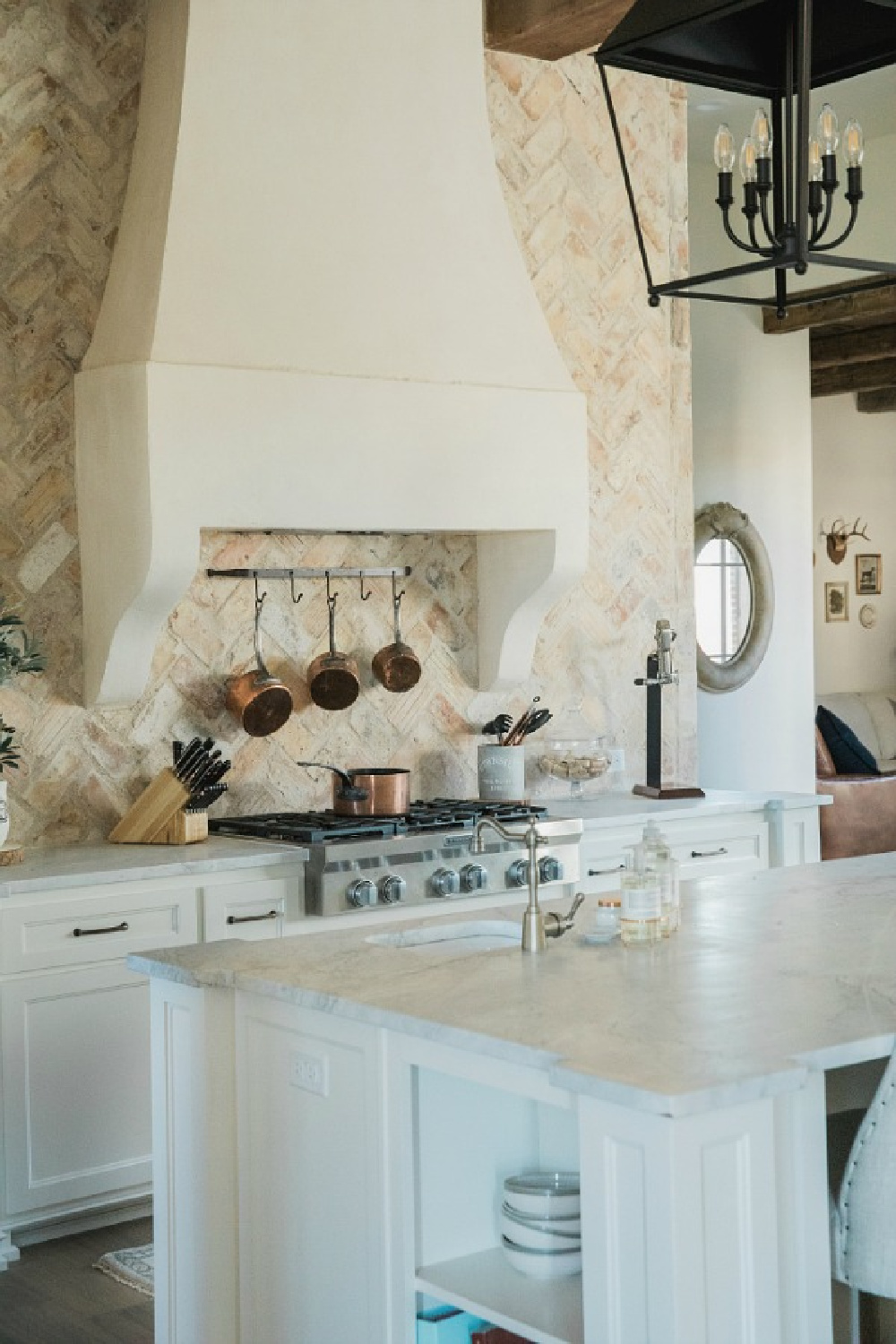 Rustic elegant French country farmhouse kitchen with beautiful stucco range hood, copper pots, reclaimed Chicago brick backsplash, arabescato marble counters, and lanterns over island. Brit Jones Design.