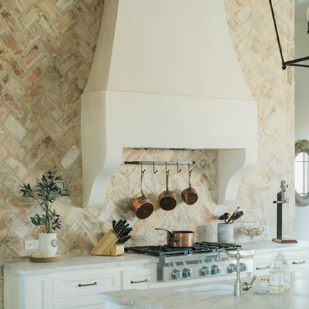 Rustic elegant French country farmhouse kitchen with beautiful stucco range hood, copper pots, reclaimed Chicago brick backsplash, arabescato marble counters, and lanterns over island. Brit Jones Design.