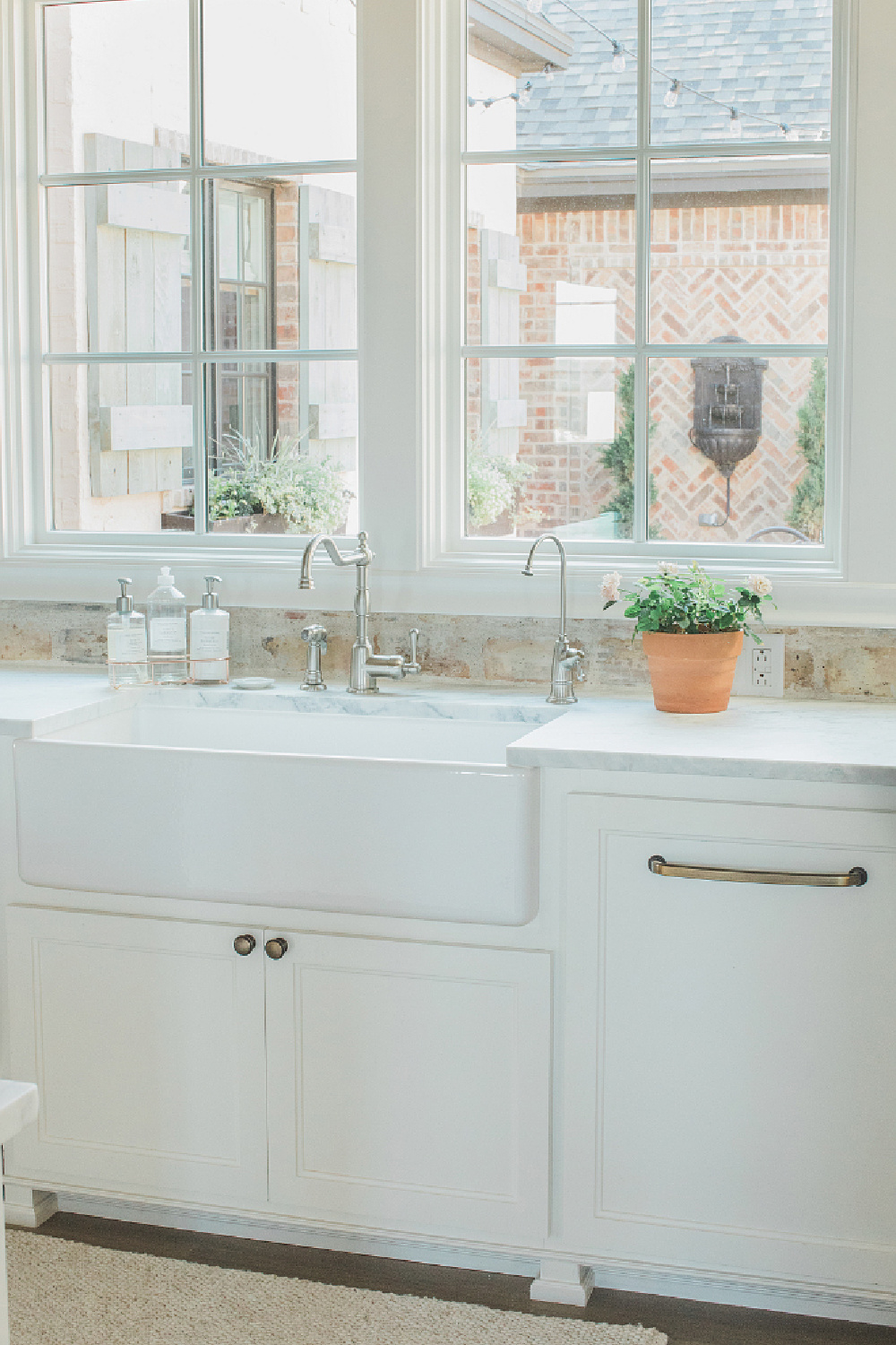 Rustic elegant French country farmhouse white kitchen with farm sink, reclaimed Chicago brick backsplash, and window overlooking courtyard. Brit Jones Design.
