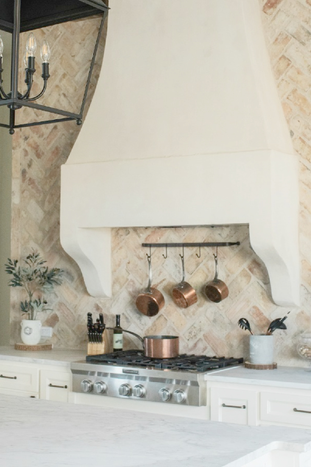 Rustic elegant French country farmhouse kitchen with beautiful stucco range hood, copper pots, reclaimed Chicago brick backsplash, arabescato marble counters, and lanterns over island. Brit Jones Design.