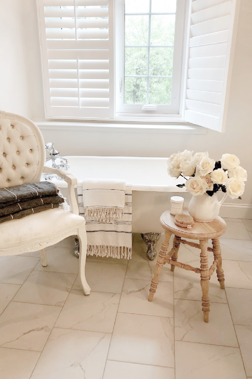 Modern French white bathroom with clawfoot tub, white roses, Louis chair, and rustic wood stool - Hello Lovely Studio.