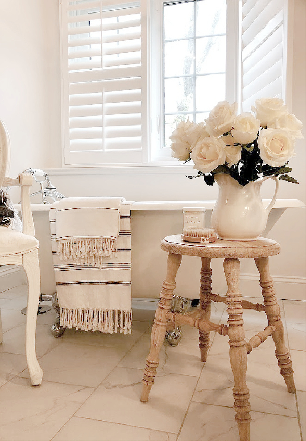 Modern French white bathroom with maid's tub, farmhous estool, white ironstone pitcher with roses, and plantation shutters - Hello Lovely Studio.