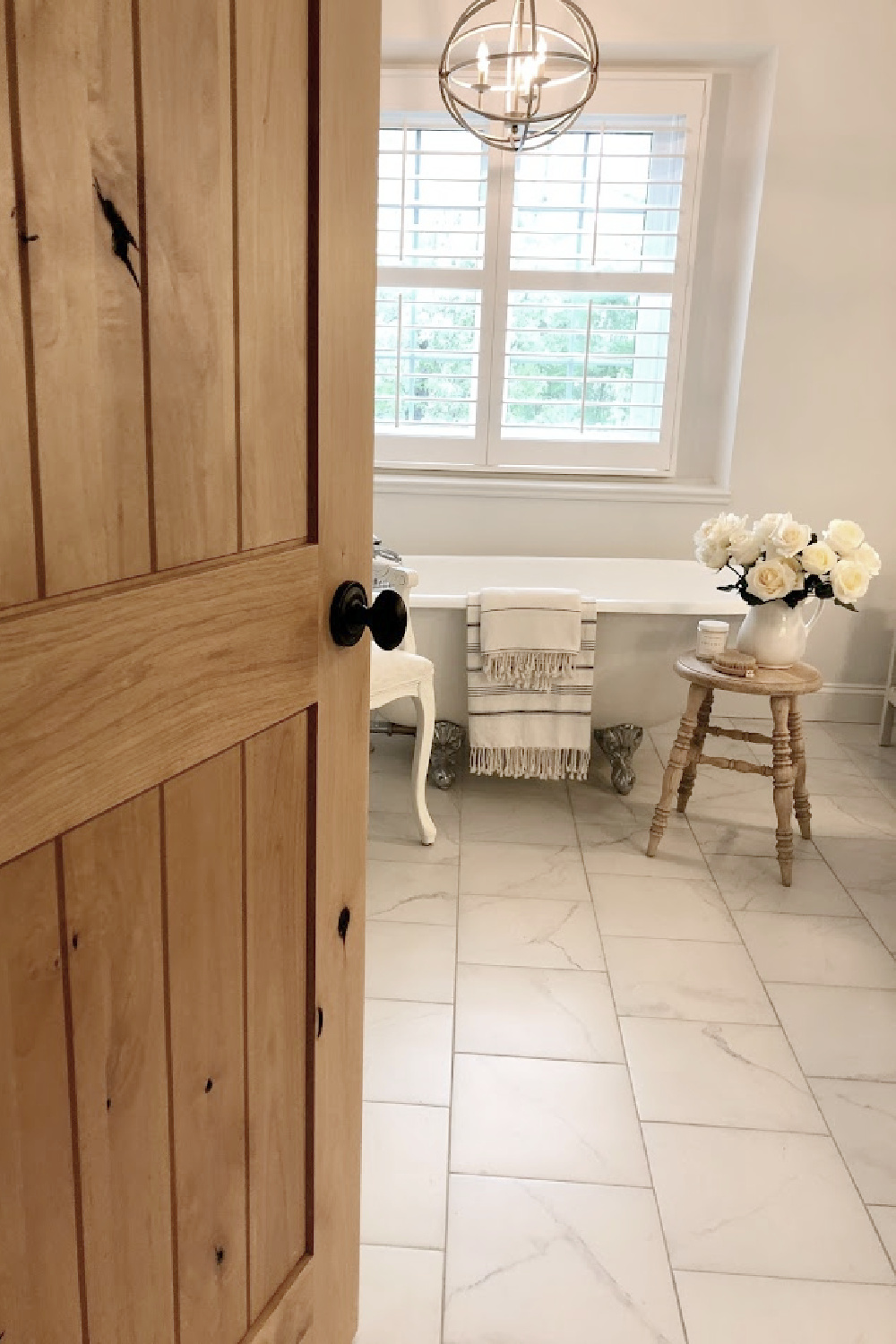 Peek inside my modern French bath with knotty alder door, clawfoot tub, white roses, and serene decor - Hello Lovely Studio.