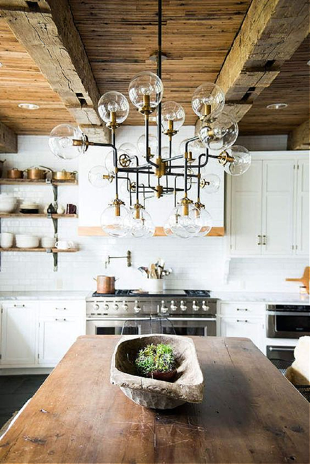 Leanne Ford rustic modern farmhouse kitchen with rugged wood ceiling beams, Sputnik chandelier over farm table, white cabinets, and stainless appliances.