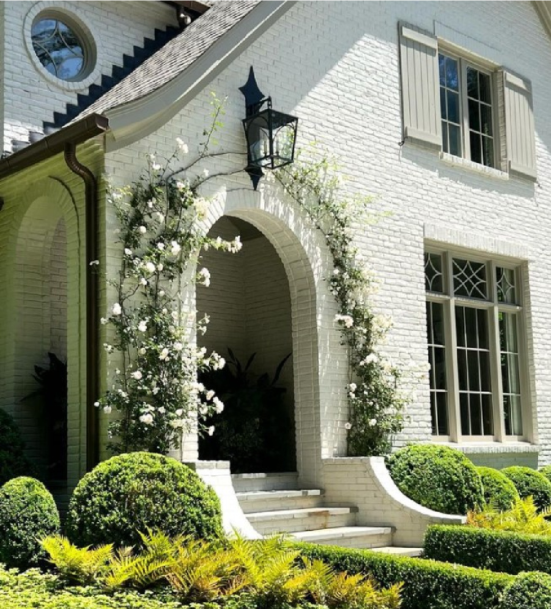 BM Balboa Mist 1549 on a brick exterior of a breathtaking storybook charm of a Tudor style white painted brick home renovated by @ladisicfinehomes with arched entrance softened by creeping roses - photo by Sherry Hart. #balboamist #bmbalboamist