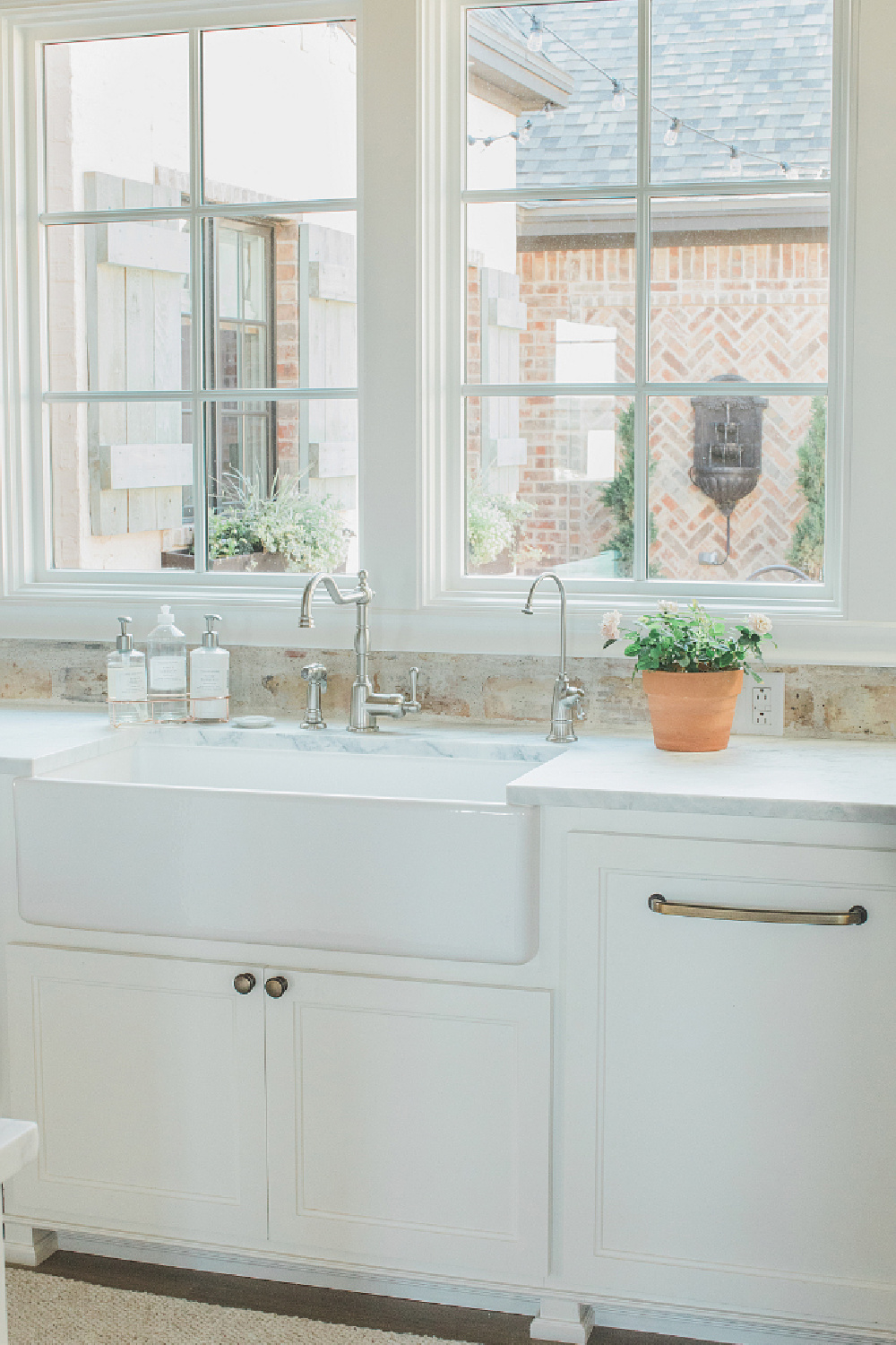 Rustic elegant French country farmhouse white kitchen with farm sink, reclaimed Chicago brick backsplash, and window overlooking courtyard. Brit Jones Design.