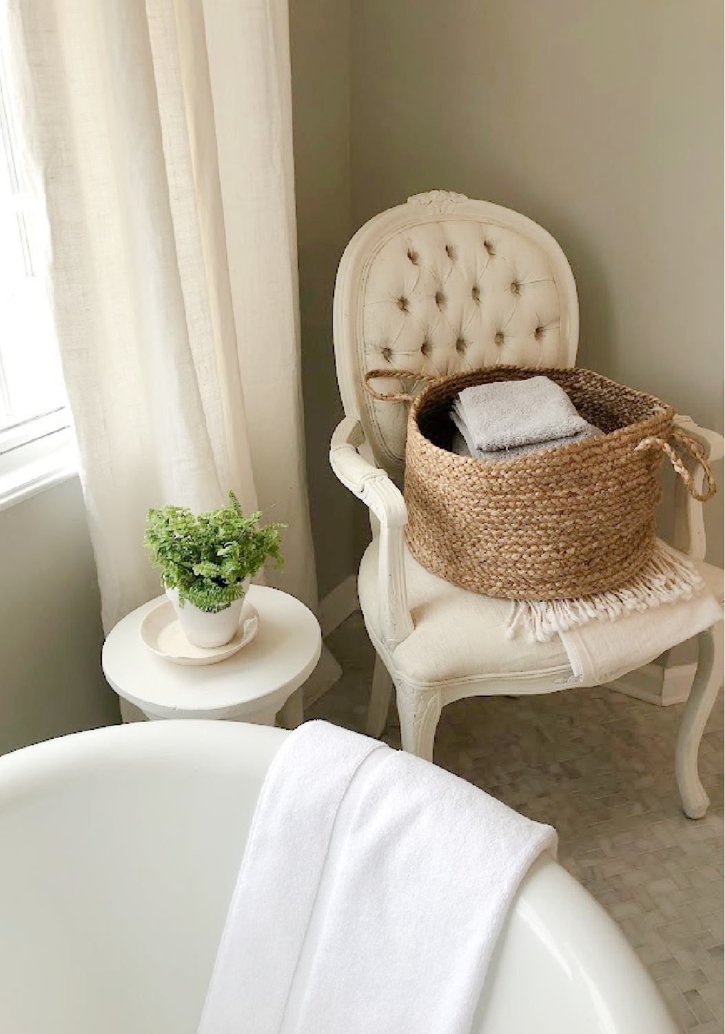 Modern French renovated bath with Louis style arm chair holding basket of towels, column side table with plant, and vintage clawfoot tub - Hello Lovely Studio. Walls are SW Repose Gray. #swreposegray
