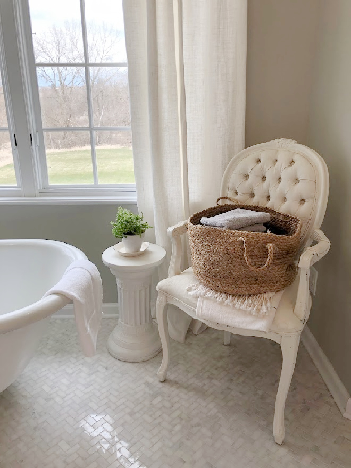 Modern French renovated bath with Louis style arm chair holding basket of towels, column side table with plant, and vintage clawfoot tub - Hello Lovely Studio. Walls are SW Repose Gray. #swreposegray