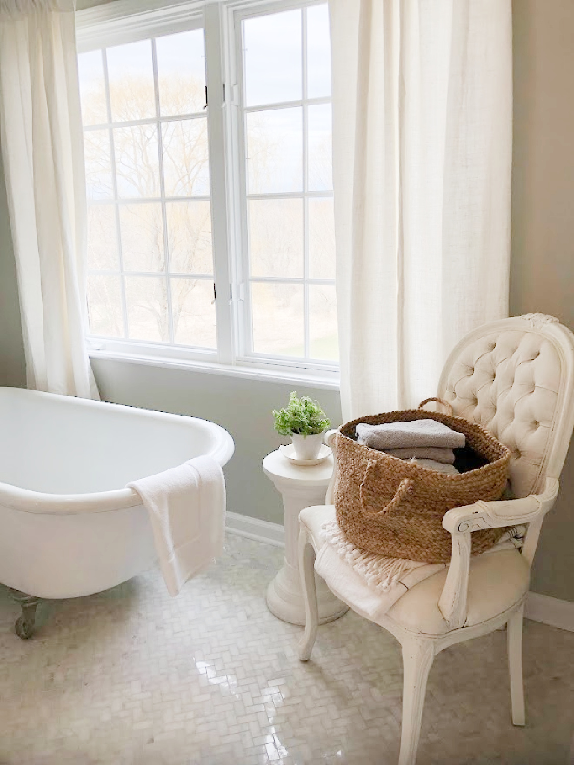 Modern French renovated bath with Louis style arm chair holding basket of towels, column side table with plant, and vintage clawfoot tub - Hello Lovely Studio. Walls are SW Repose Gray. #swreposegray