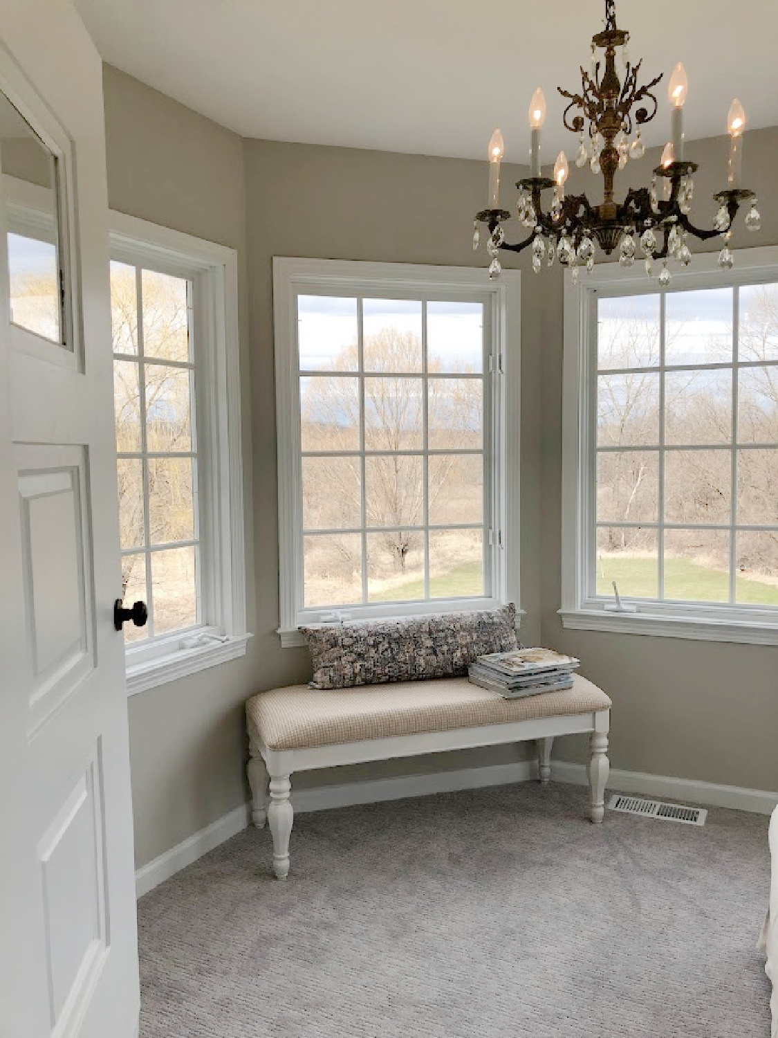 French country bench, antique brass chandelier, and undressed windows in turret sitting room of coastal bedroom remodel (SW Agreeable Gray on walls) at the Georgian - Hello Lovely Studio. #agreeablegray #swagreeablegray