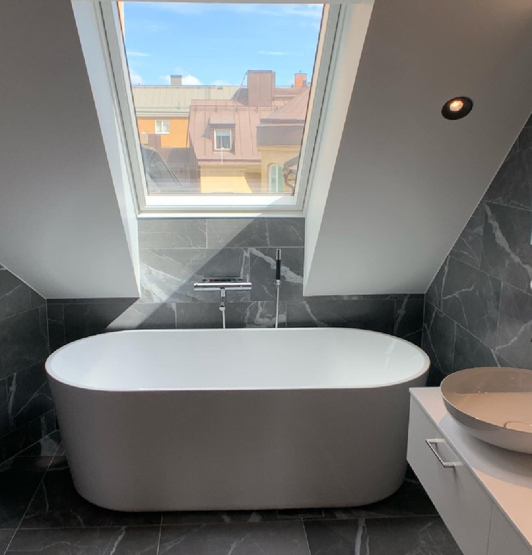 Modern soaking tub with a view of city in dark grey tiled bath in a Stockholm apartment - @residensportalen. #stockholmapartment #modernbathroom