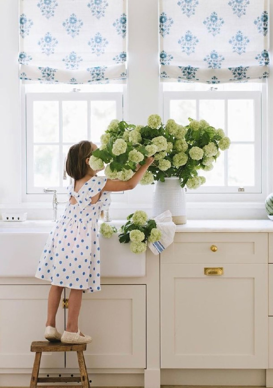Schumacher 1889 fabric on roman shades in a beautiful white kitchen with farm sink, little girl in polka dot dress, and vase of limelight hydrangeas. #blueandwhitekitchen