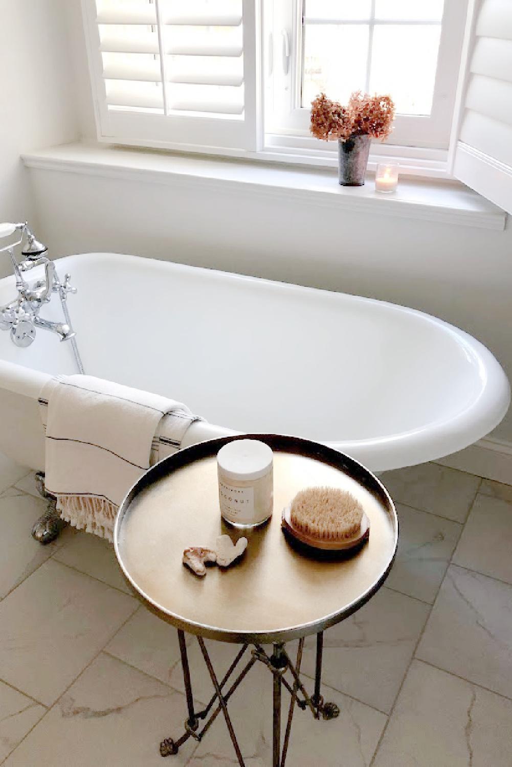 My serene bath with small clawfoot Maid's tub, plantation shutters, and round silver accent table - Hello Lovely Studio. #serenebath #bathroomdesign #minimalluxe
