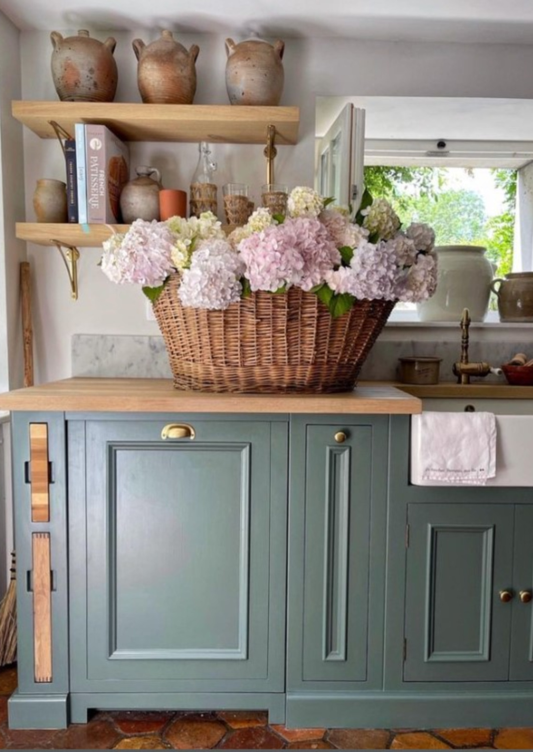 French farmhouse kitchen in France with green cabinets (Farrow & Ball Green Smoke), butcher block counters, and basket of pink hydrangea - Vivi et Margot. #vivietmargot #frenchfarmhouse #greenkitchens