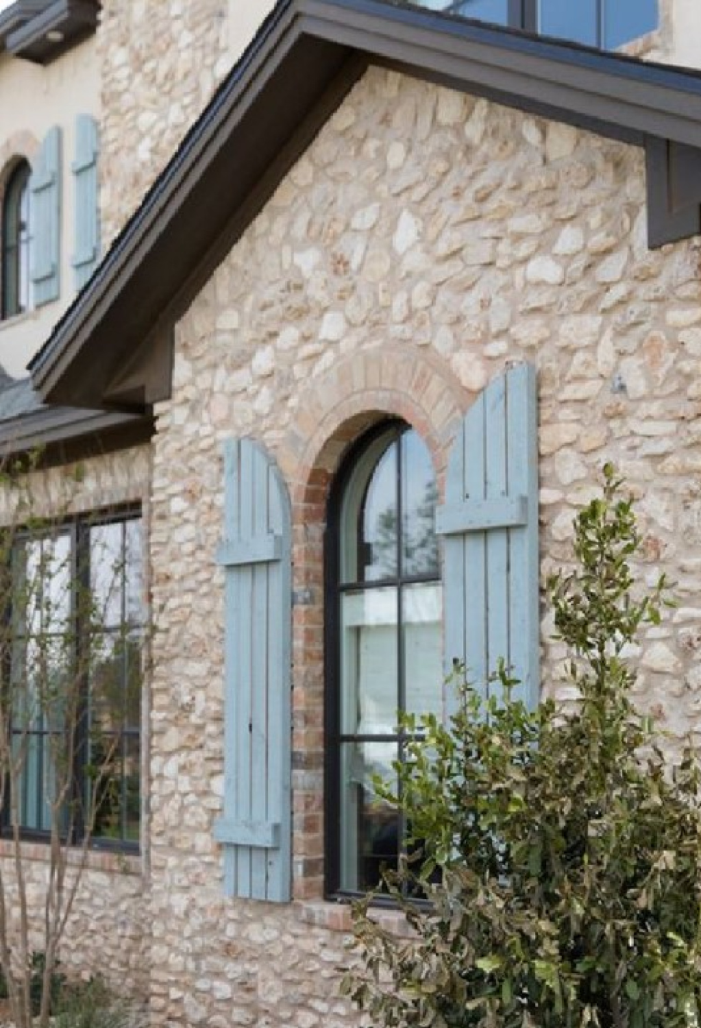 Modern French house exterior with warm stone, dark brown trim, arched windows, and duck egg or French blue rustic shutters. Brit Jones Design. #modernfrench #frenchcountryarchitecture