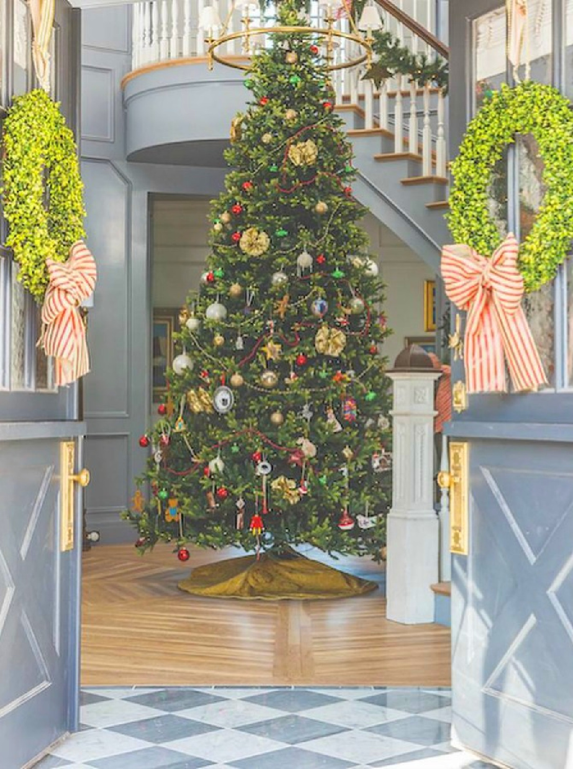 Holiday decorated front doors with boxwood wreaths and striped ribbons and foyer (with blue grey painted moldings) with grand Christmas tree at magnificent staircase - The Fox Group. #christmasfoyer #elegantchristmas