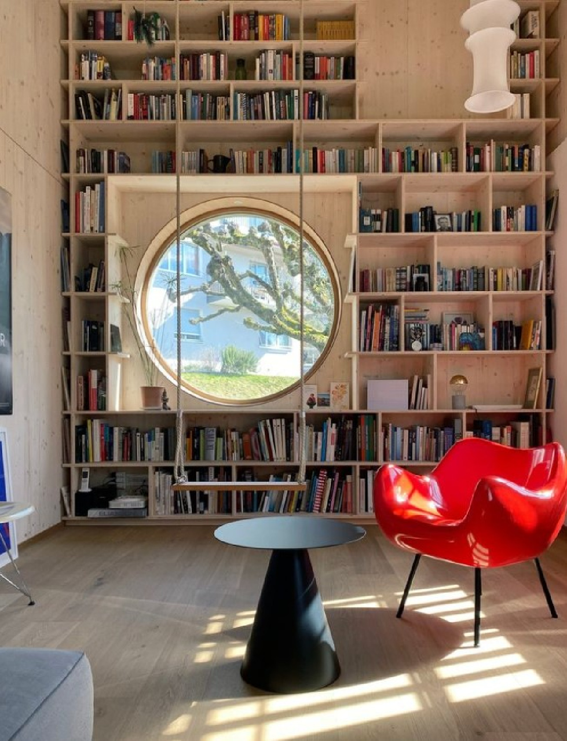 Modern home library with round window incorporated in bookshelves and sculptural red chair - The World of Interiors.